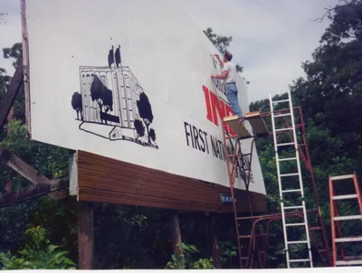 Jim Ford stands on scaffolding, painting an advertisement onto a roadside billboard.