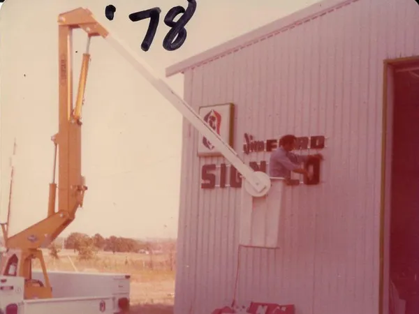 A man in a crane basket puts the finishing touches on a sign mounted on a large, metal work shed.